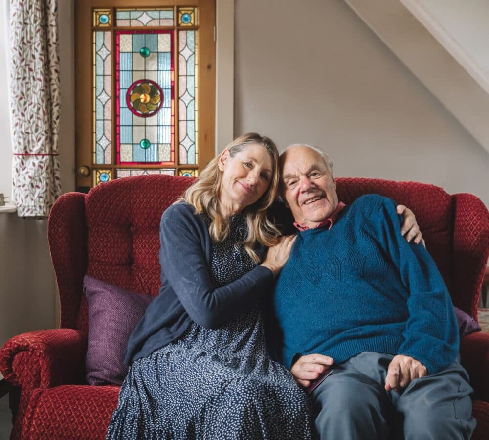 Older man with his daughter sitting both on a couch inside their home both happy and smiling