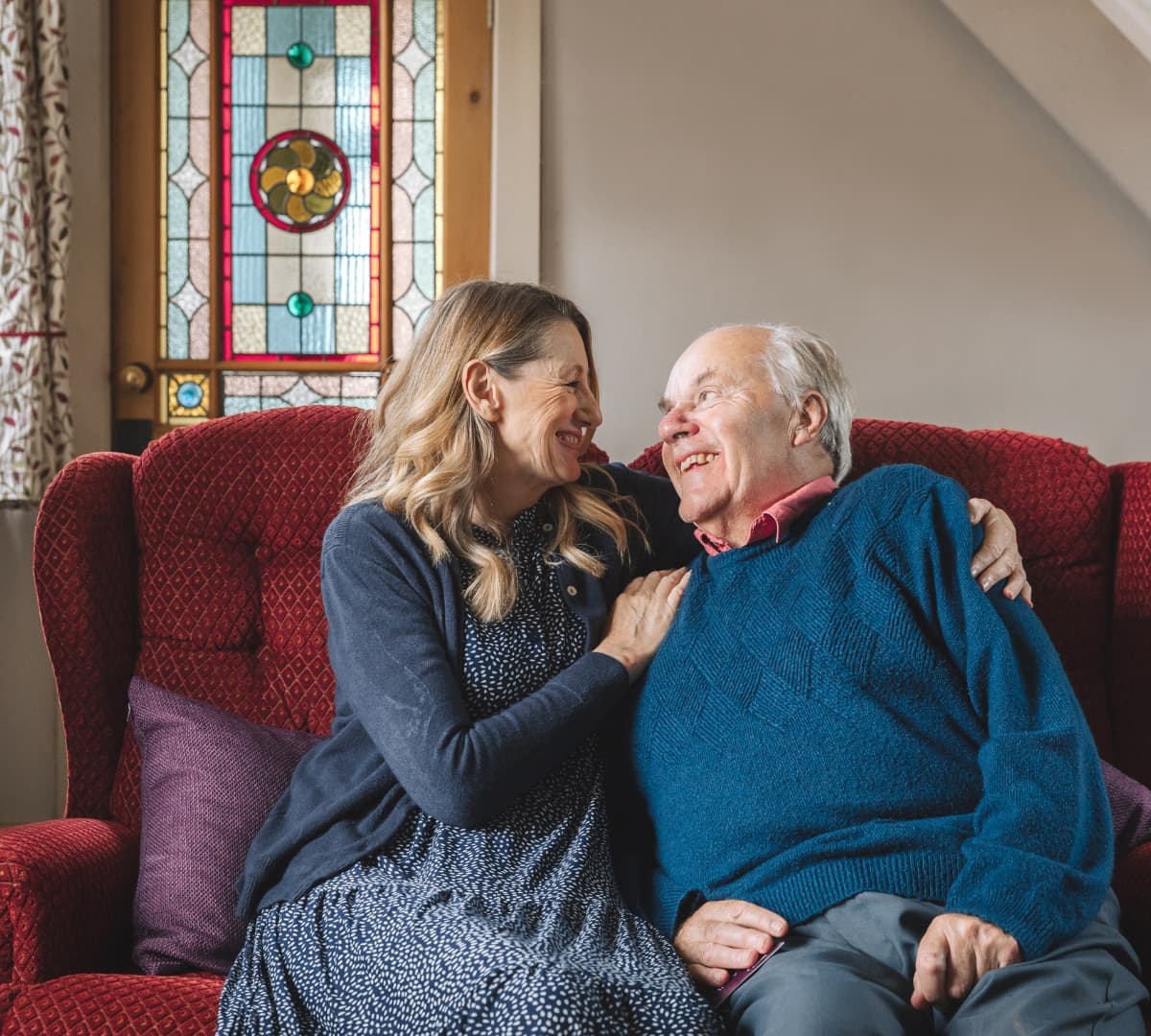 Older man with white hair sitting on a red couch with his daughter with long blonde hair smiling and looking at each other inside the house