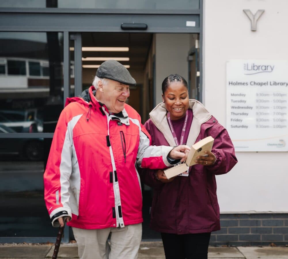 Older man wearing a jacket and a hat going out of the library with his female carer while looking at a book both smiling and happy