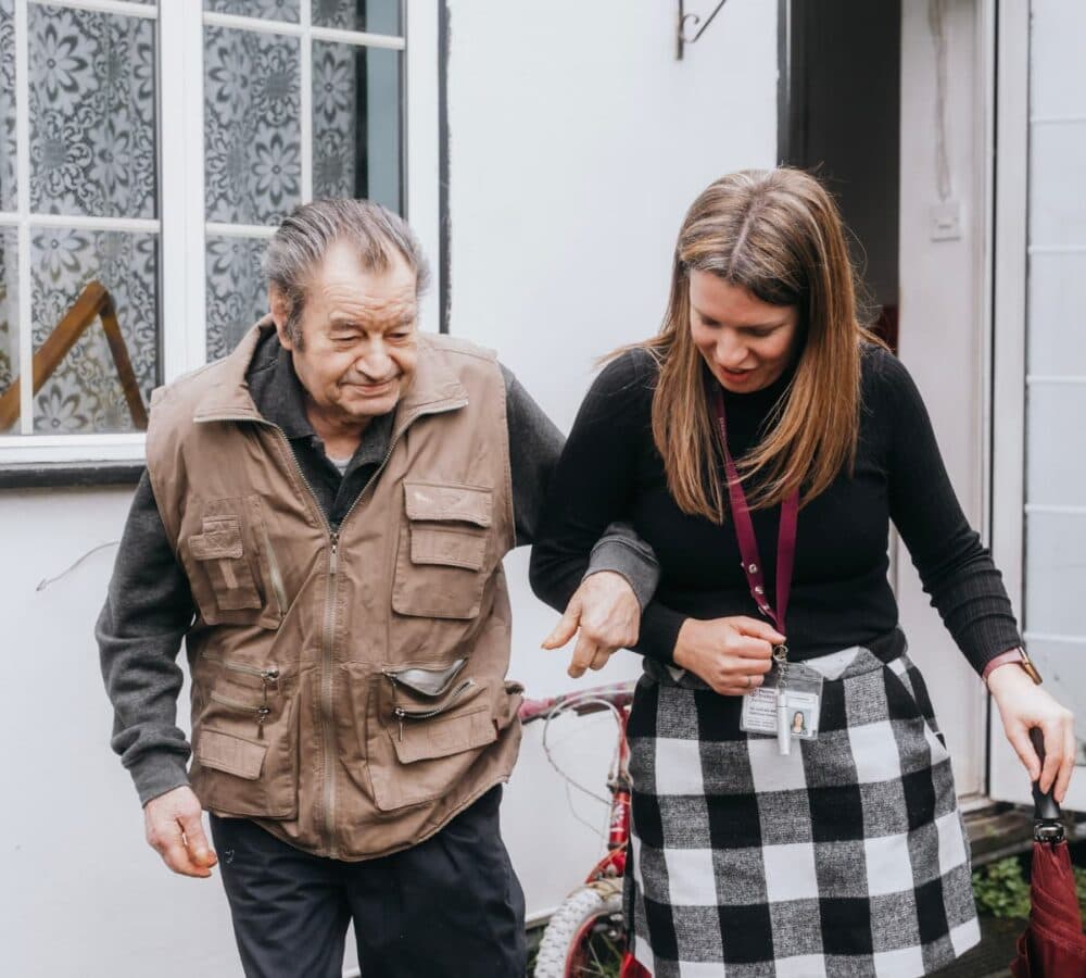 A woman supports an elderly man as they walk together outside a house. - Home Instead
