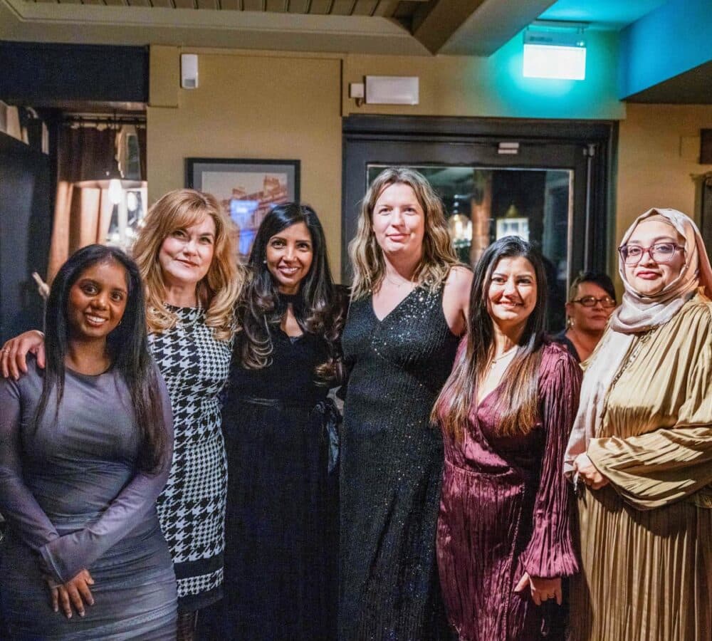 Six women in formal attire smile and pose together indoors at an evening event. - Home Instead