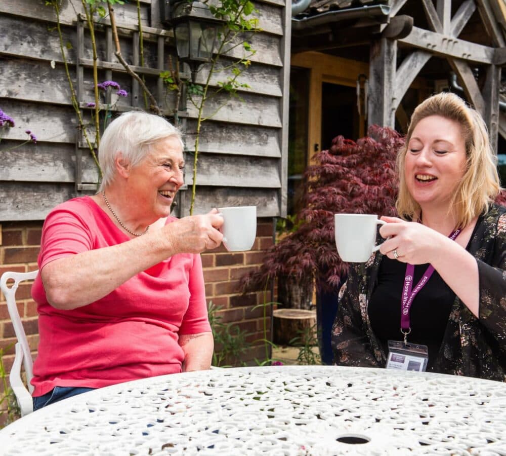 An older woman having coffee while toasting with her carer outside and both are happy
