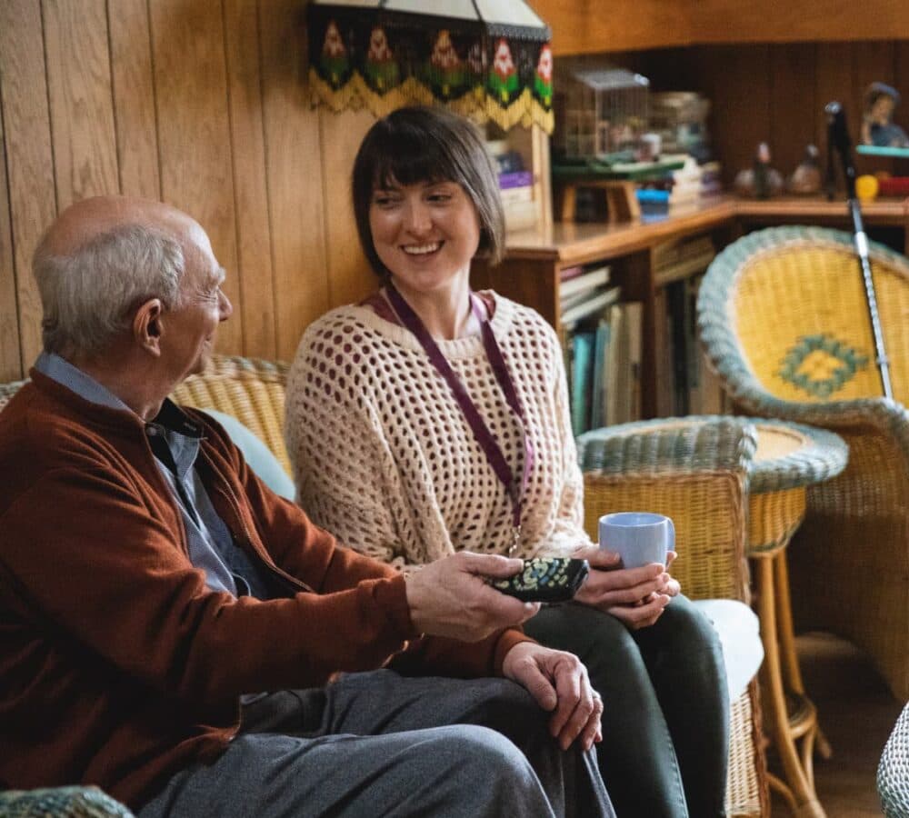An older man sitting on a couch with his younger female carer with short long black hair both happy and smiling inside the home