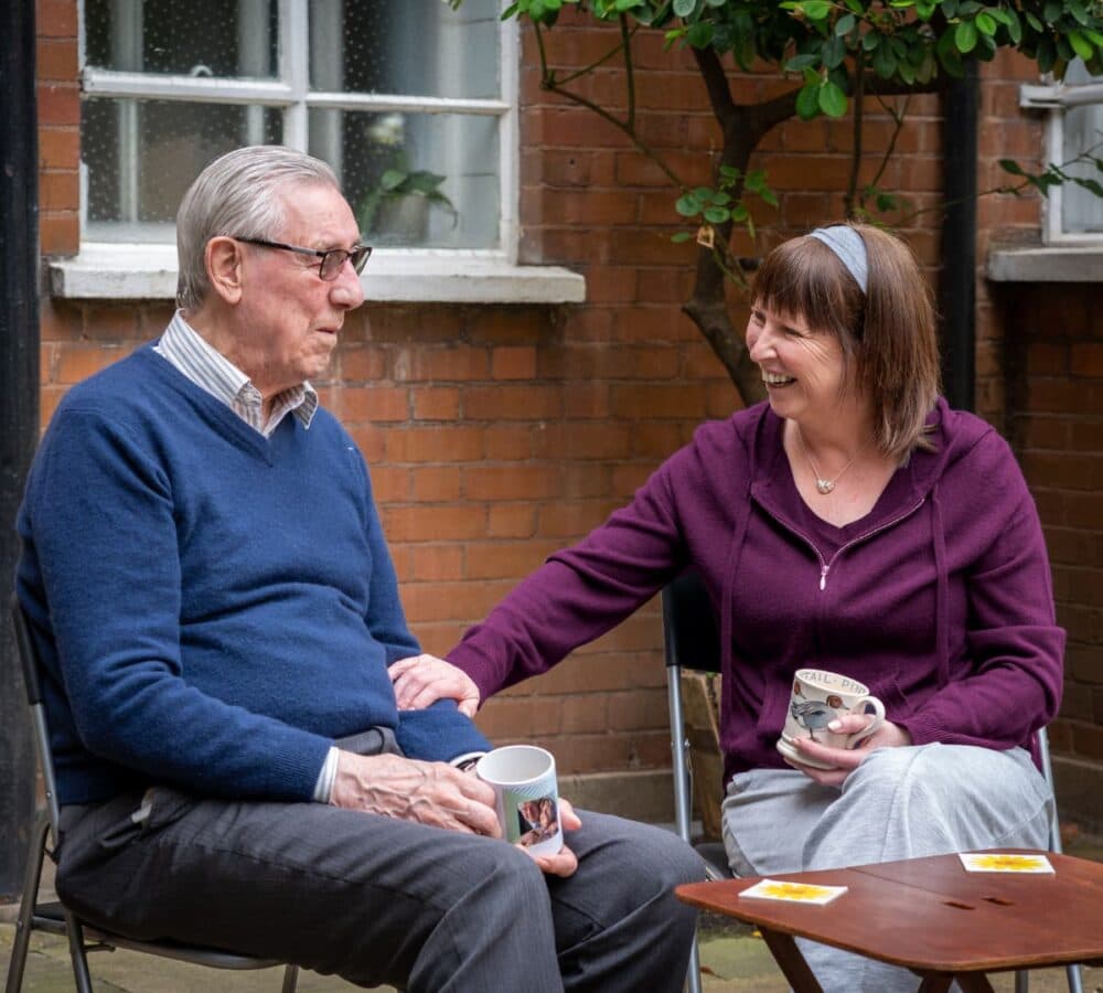 An older man wearing blue sweater and with grey hair and eyeglasses having coffe outside the house with his younger female carer with short hair and both happy while chatting