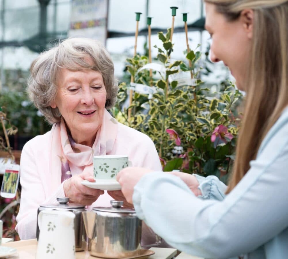 Older woman with grey hair having a cup of tea with her younger female carer outdoor
