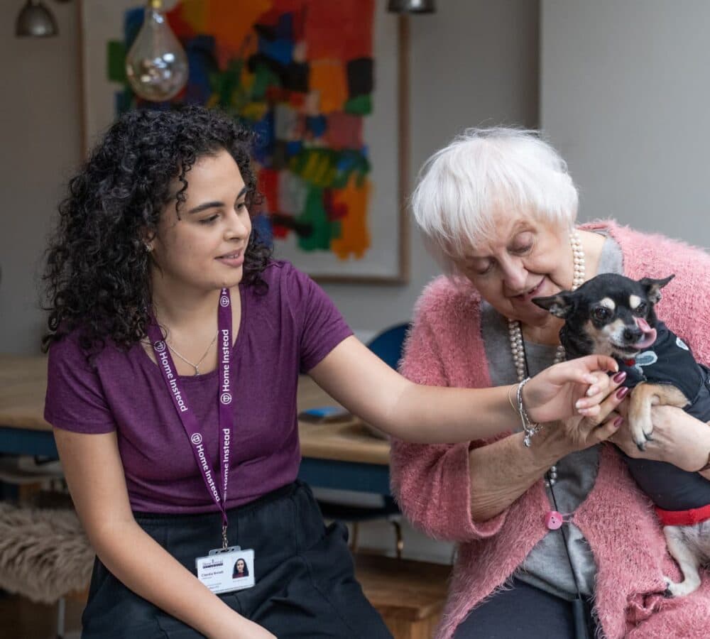 Older female woman with white hair and wearing pink sitting inside the house while holding her dog with her younger carer with long blonde hair and wearing purple shirt