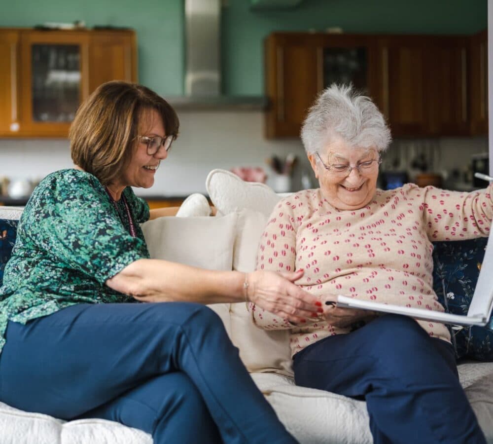 An older woman looking at a photo album while sitting on a couch with her younger carer with short hair and wearing green