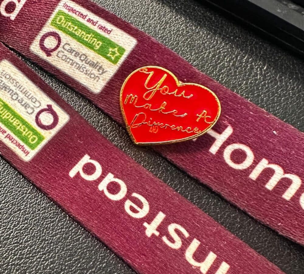 A red heart pin reading "You make a difference" on a maroon Homestead lanyard with Care Quality badges. - Home Instead