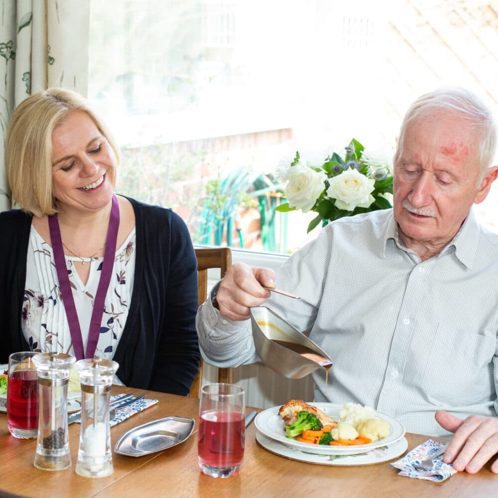 Elderly man having lunch with his Home Instead carer, showing him pouring gravy whilst the carer sits next to him.