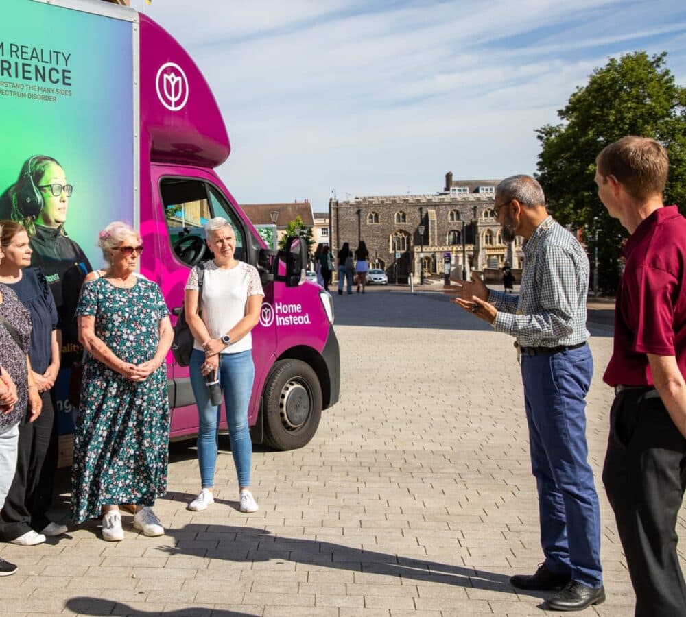 A man talking to a group of women outdoor with sunshine and a dementia bus at the side