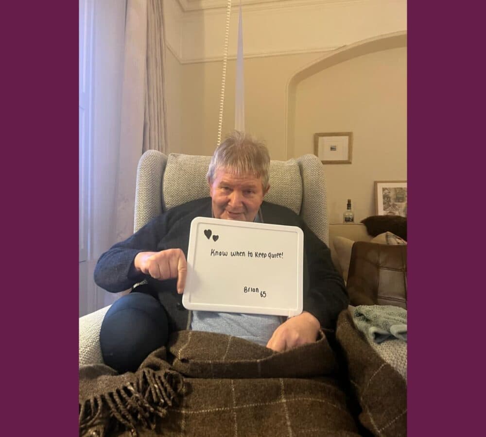 Older man, brian with grey hair sitting on a couch while holding a sign that says Know when to keep quiet.