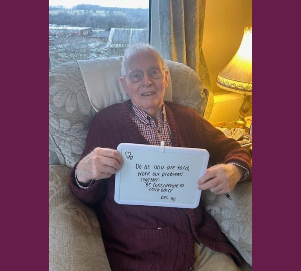 Older man with grey hair sitting on a couch near the window and wearing a sweater while holding a sign board
