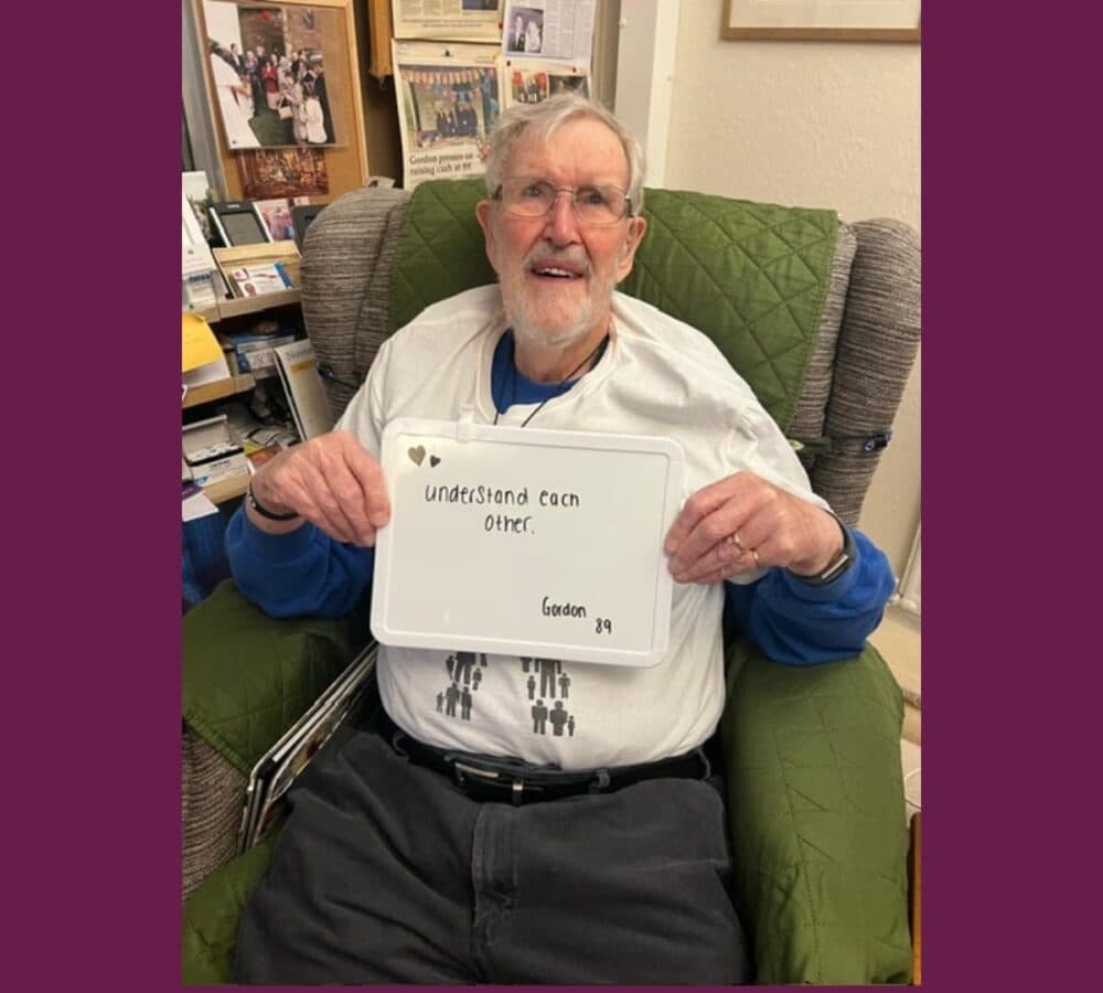 Older man Gordon with white hair sitting on a green couch while holding a sign that says Understand each other
