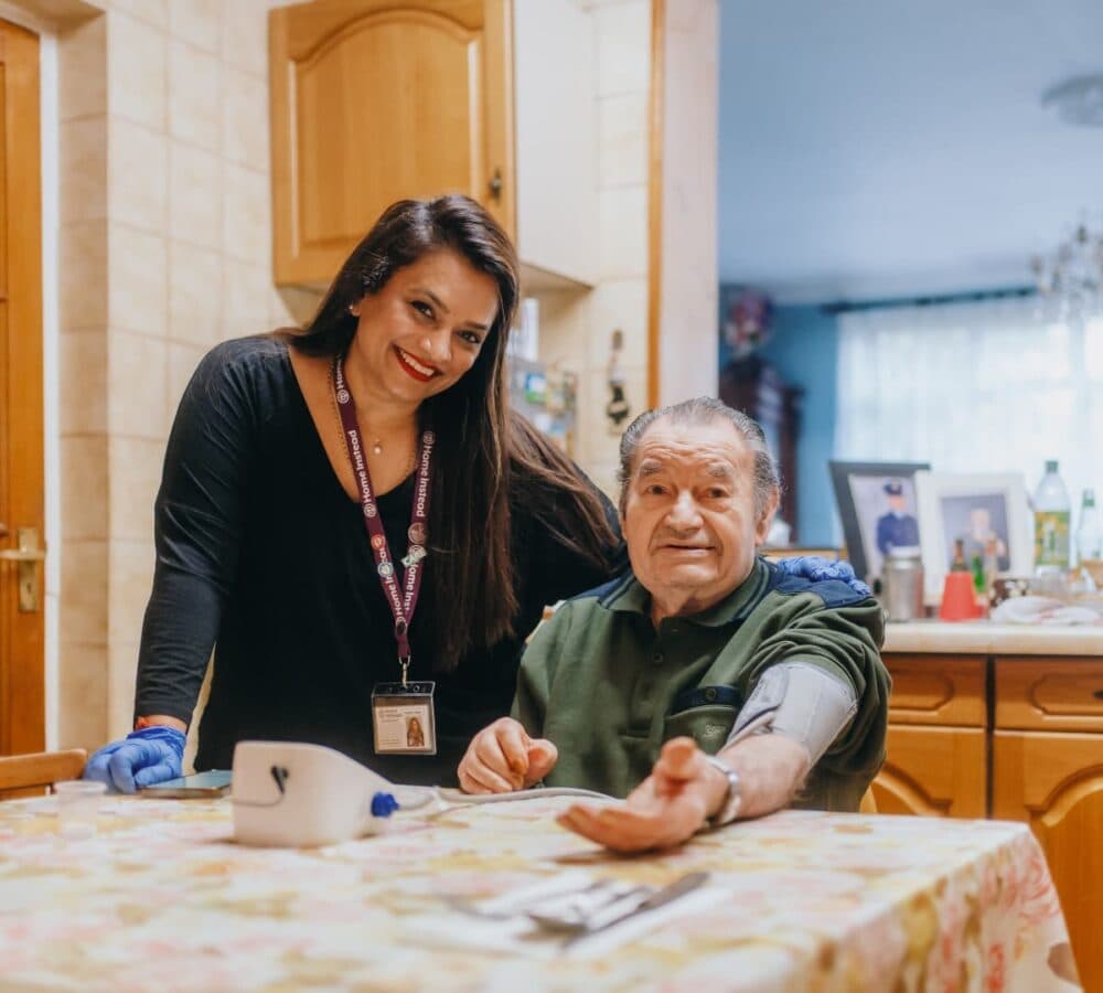 Older man sitting on a chair and his arm on top of the table with his younger female carer with long black hair and wearing gloves inside the kitchen and both smiling