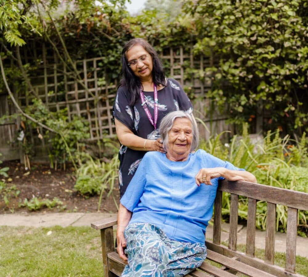 Older adult woman with grey hair and wearing blue and sitting on a bench with her female carer with long black hair surrounded by plants in the garden