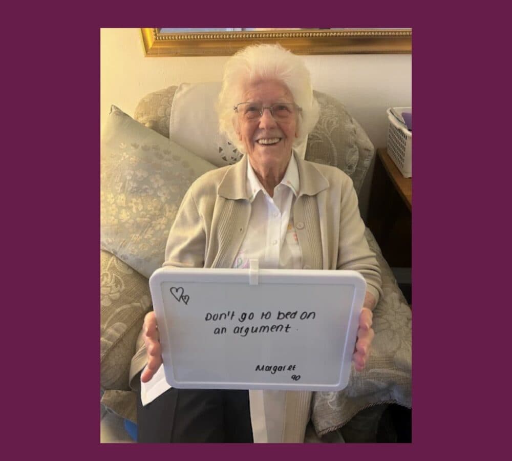 Older woman named Margaret with white hair sitting on a couch while holding a sign saying Don't go to bed on an argument