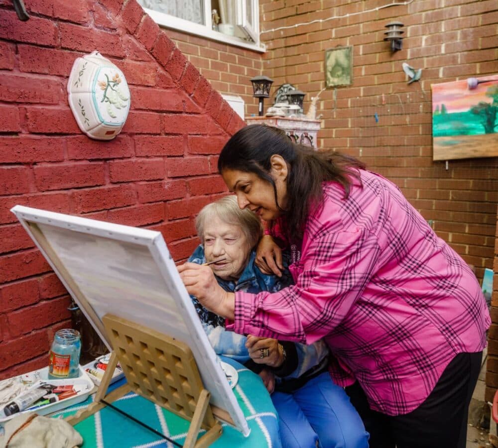 Older woman with short hair outside the house while painting with her carer with long black hair and wearing pink