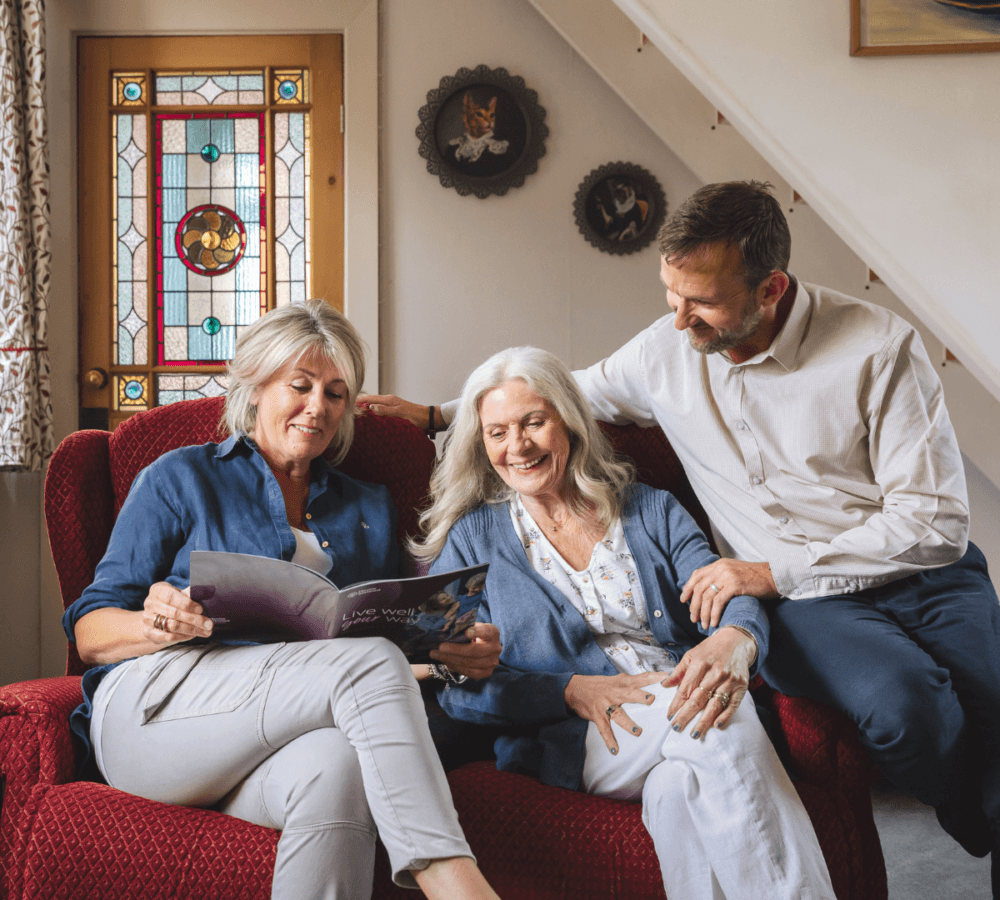 older woman with long grey hair talking to her son and daughter all smiling while sitting on a couch and looking at a Home Instead brochure