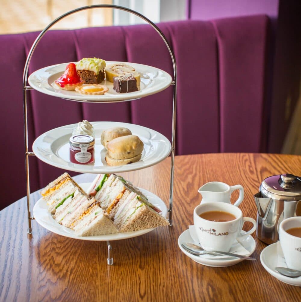 Three-tiered afternoon tea stand with sandwiches, scones, desserts, and a cup of tea on a wooden table. - Home Instead