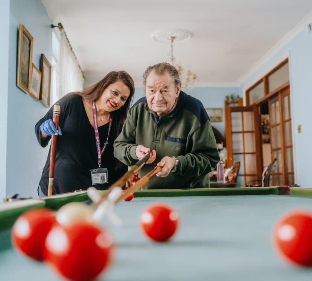 A male older adult with grey hair playing billiard with his younger female carer with long black hair and wearing gloves inside the house