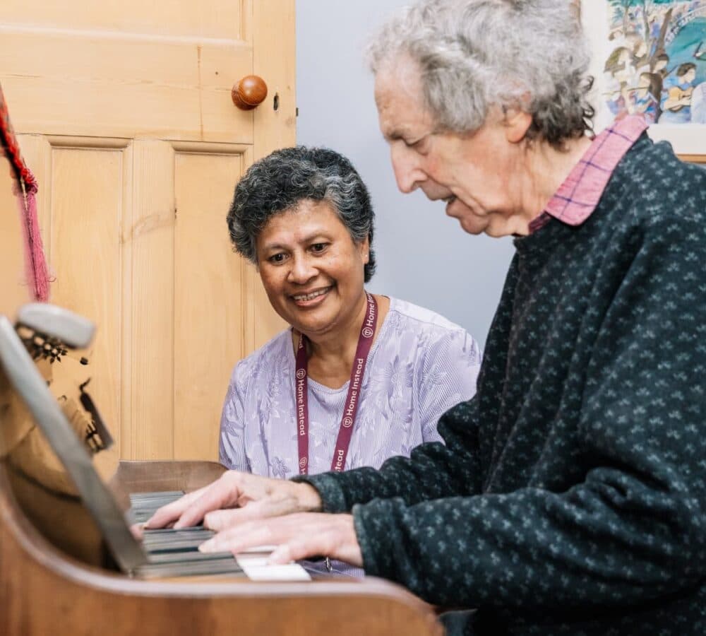 Older man with grey hair playing the piano with his female carer happily watching him