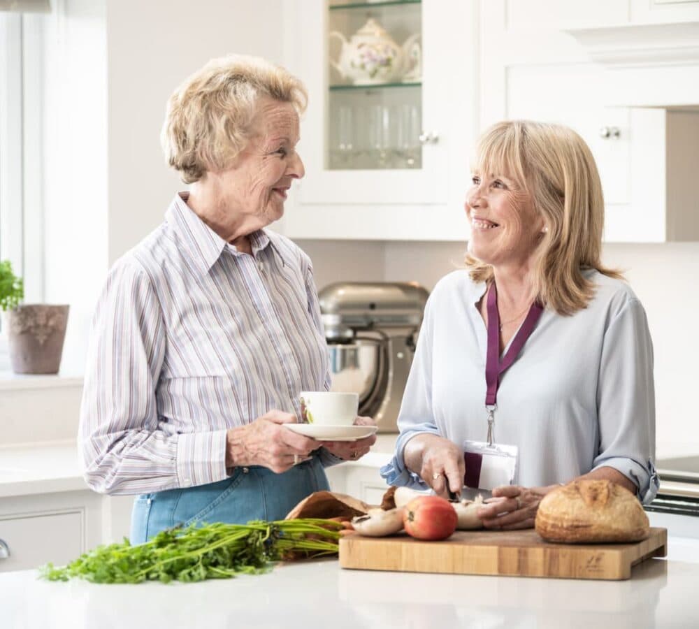 Female carer with short hair chopping foods in the kitchen while chatting with a female older adult who is drinking coffee