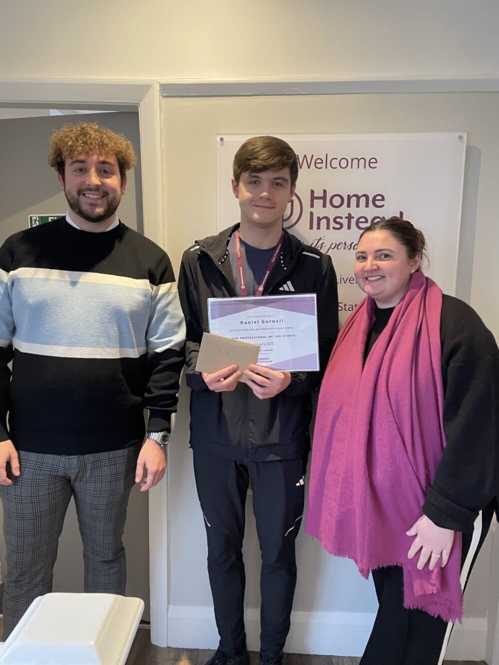 Three people smiling indoors, with the center person holding a certificate and envelopes. - Home Instead