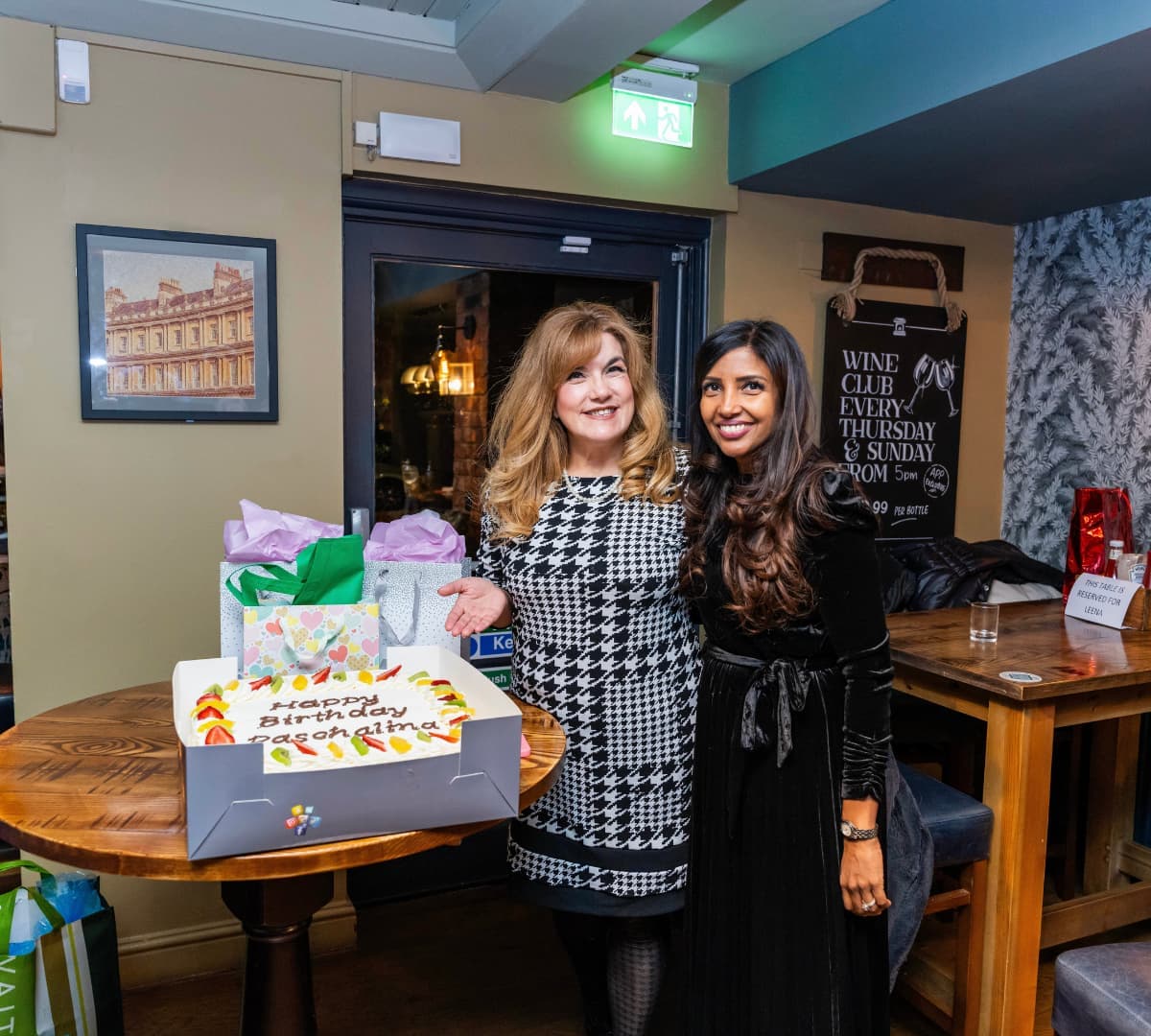 A woman wearing black dress happy and smiling with long hair and another woman with long blonde hair happy and smiling with cakes and gifts inside the restaurant