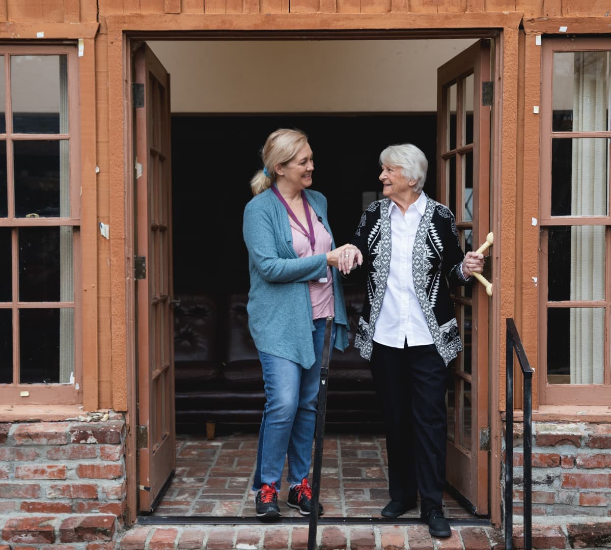 Older woman with grey hair walking out the door while being helped by her younger female carer