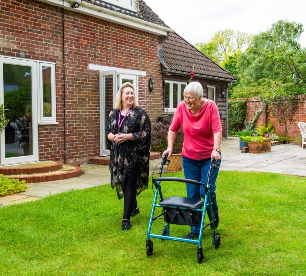 older female adult walking outdoor while using a walker with her younger female carer both smiling and surrounder by greeneries