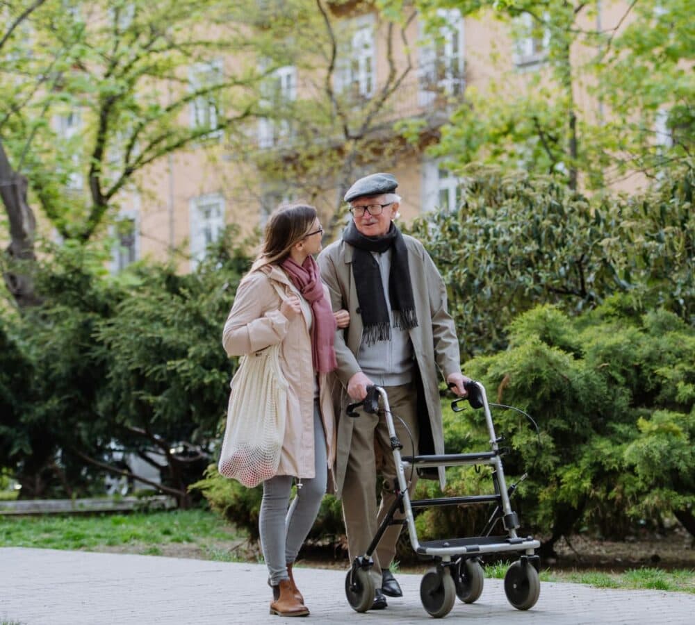 An older woman walking outdoors using a walker with his young female Care professional assisting him both smiling and happy and surrounded by greeneries