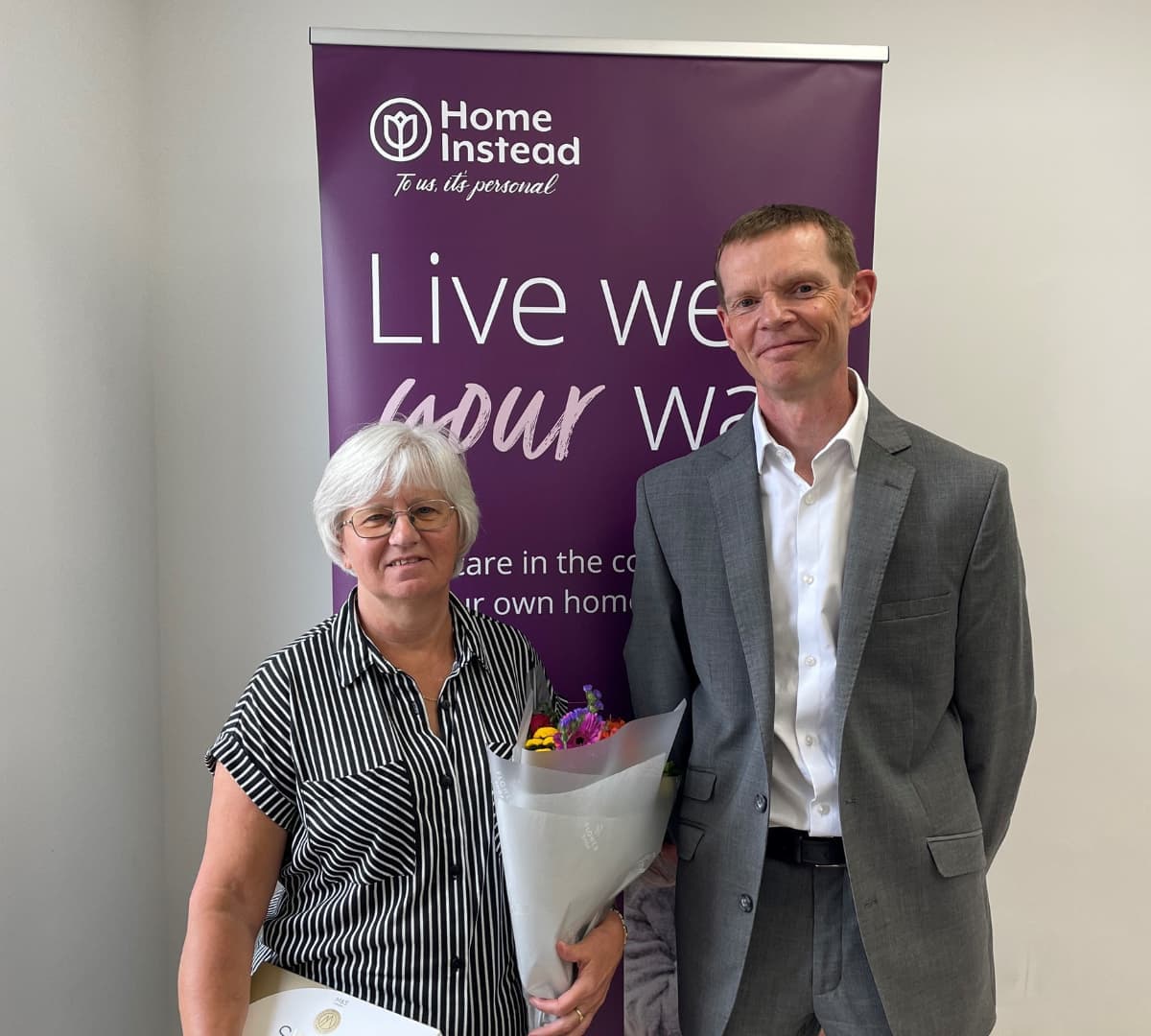A man with back hair smiling with a woman shorter than him smiling and holding a bouquet of flowers with a Home Instead banner at the back
