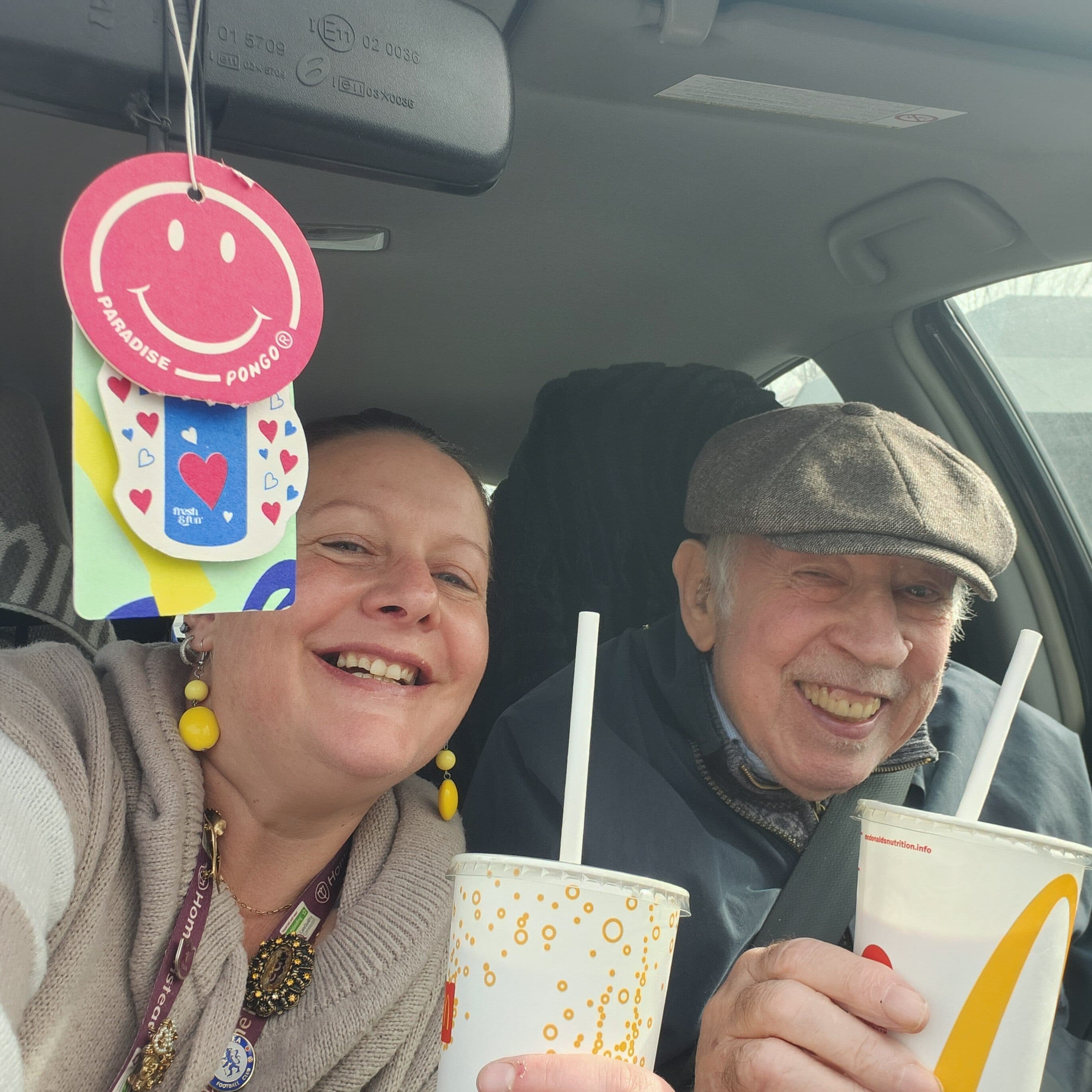 Smiling woman and elderly man in a car, holding drinks, with a red smiley air freshener hanging above. - Home Instead