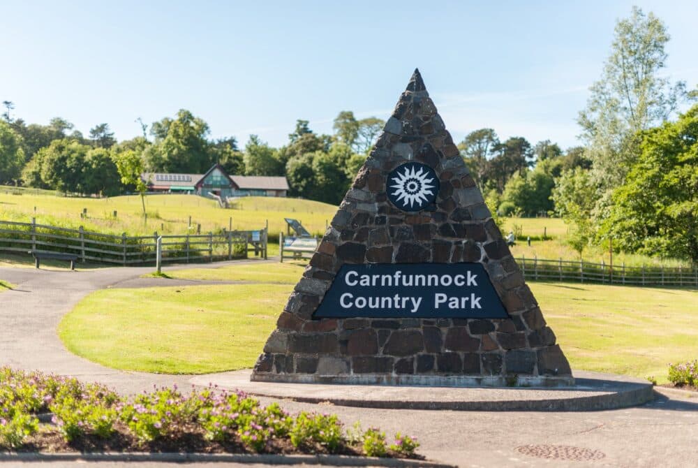A stone pyramid sign reads "Carnfunnock Country Park" with green fields and trees in the background. - Home Instead