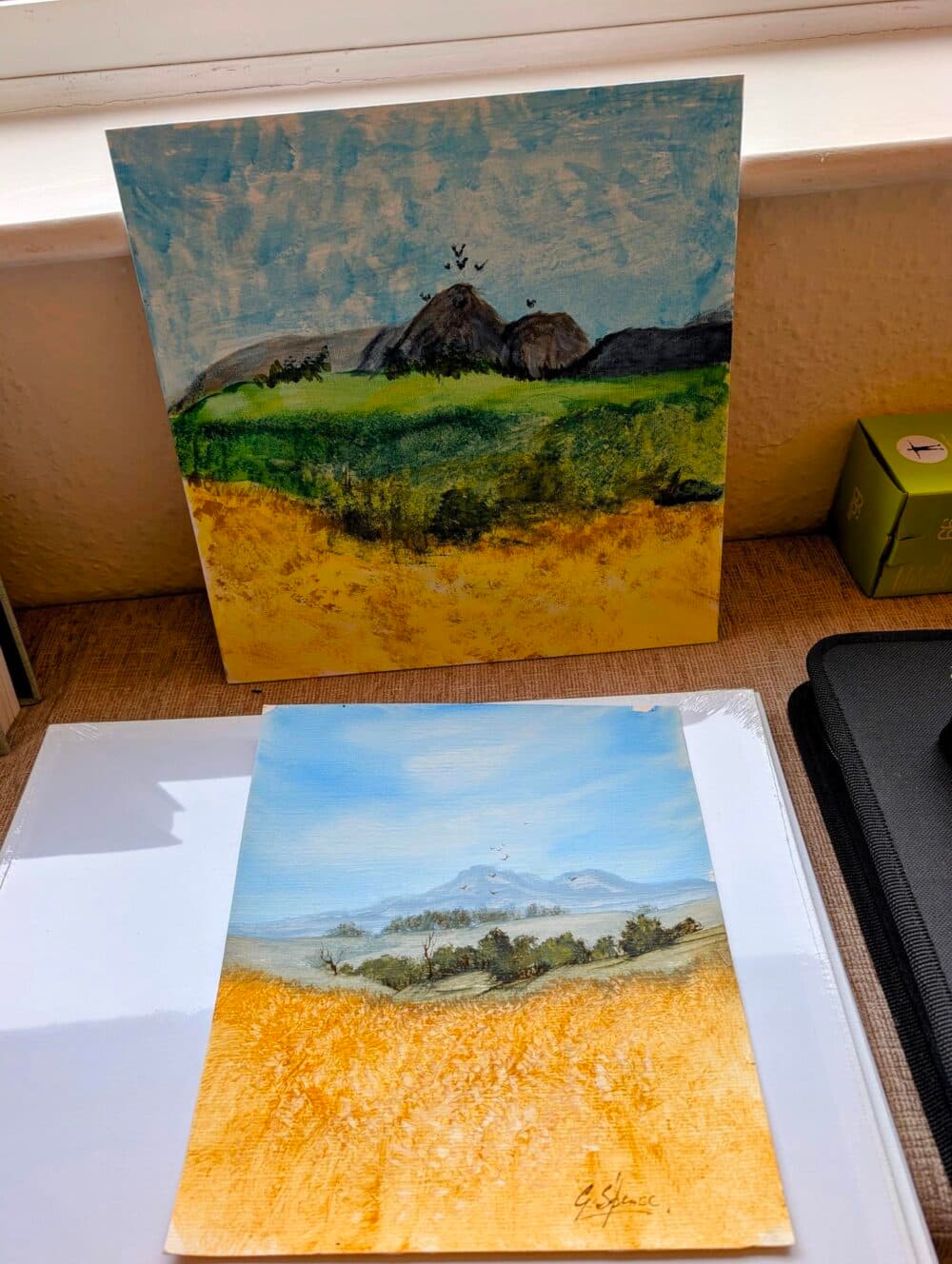 Two landscape paintings with green hills, blue skies, and yellow fields displayed on a table by a window. - Home Instead