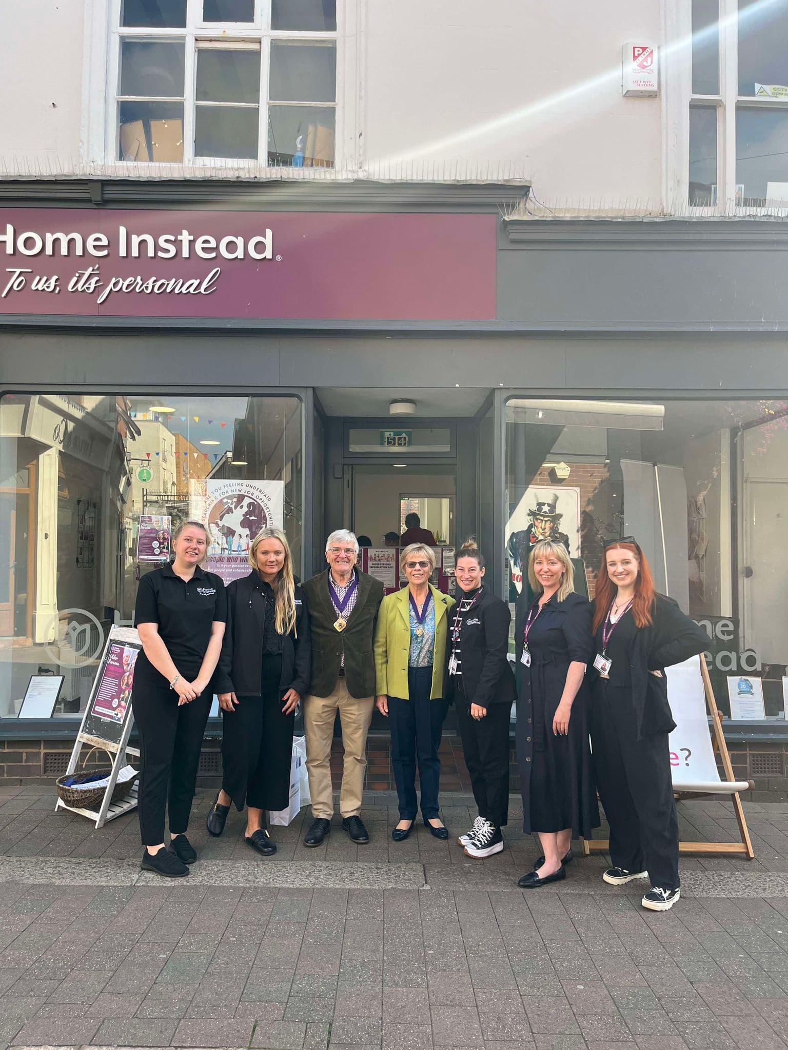 Seven people standing and smiling in front of a Home Instead storefront on a sunny day. - Home Instead