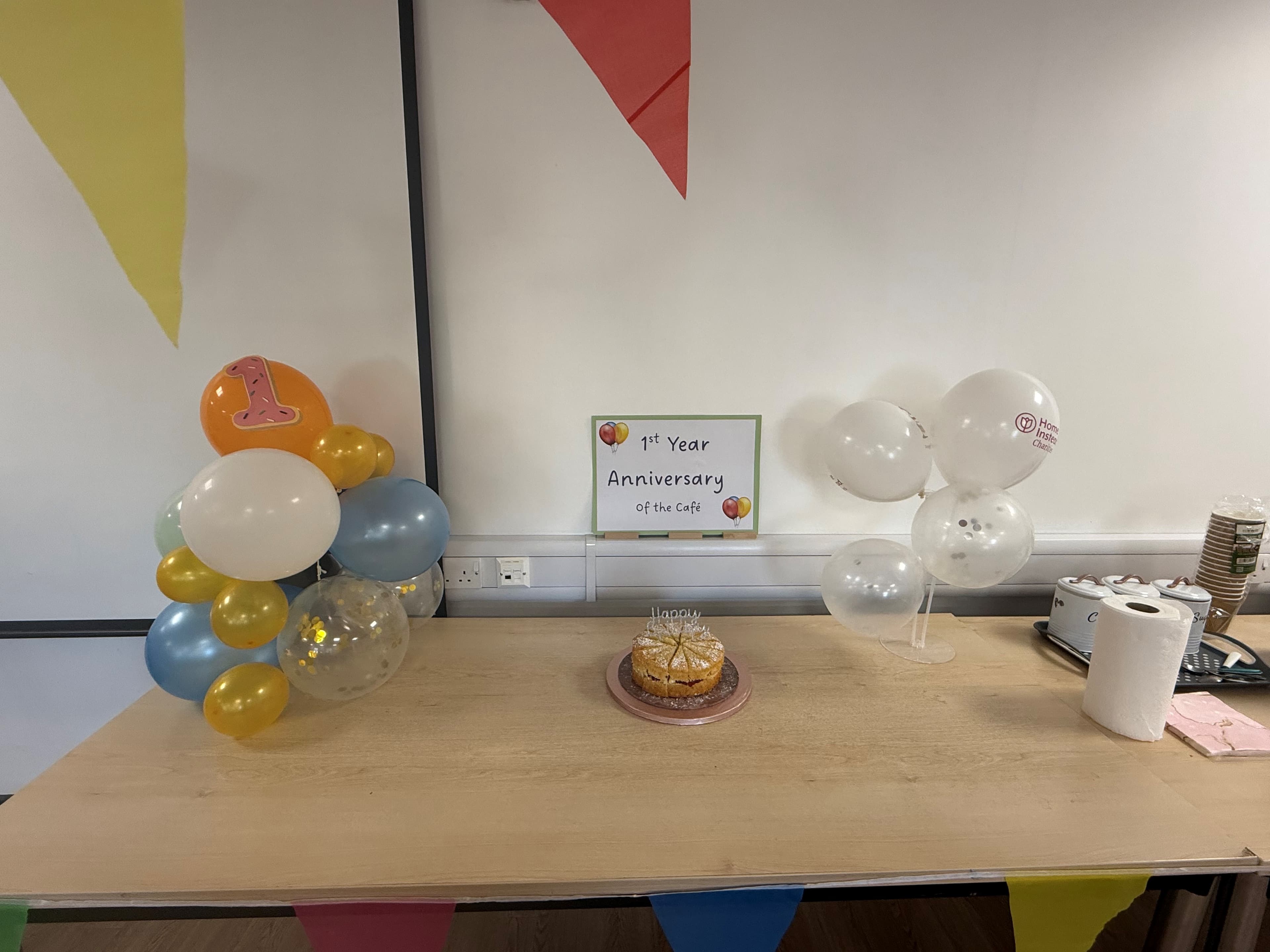 Table with balloons, a cake, and a sign reading "1st Year Anniversary of the Café" against a decorated wall. - Home Instead