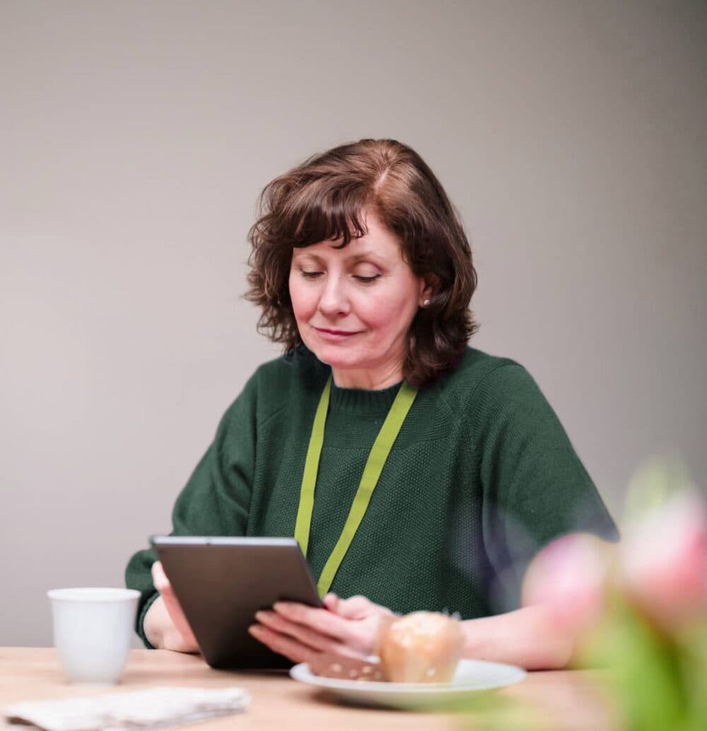 Woman with brown hair reads a tablet at a table with a coffee cup and pastry in front of her. - Home Instead