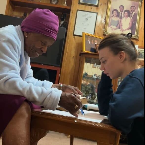 Elderly woman and young woman sit at a table, working together on papers in a cozy room. - Home Instead