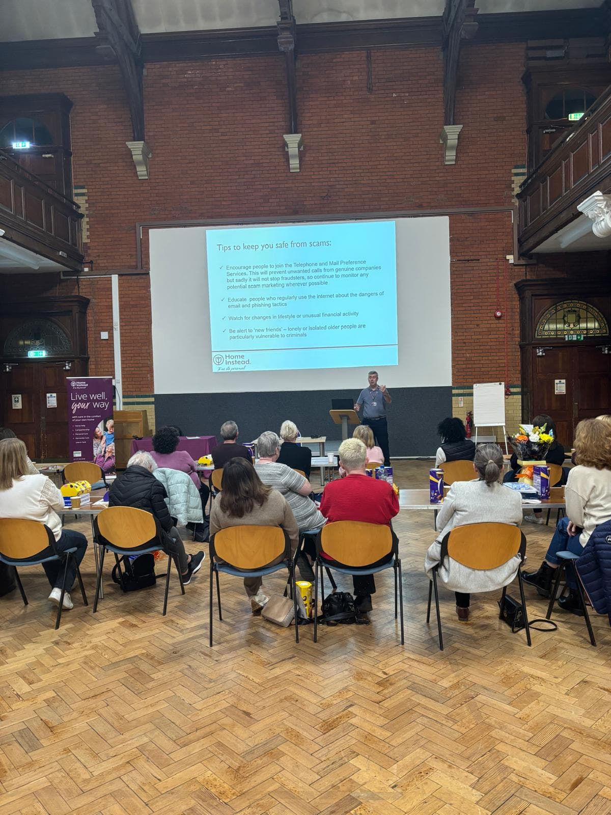A speaker presents scam safety tips to an audience in a large hall with a projected slide behind them. - Home Instead