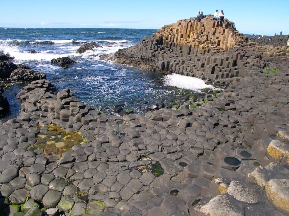 People sit atop Giant’s Causeway, surrounded by hexagonal basalt columns and waves along the rocky shore. - Home Instead