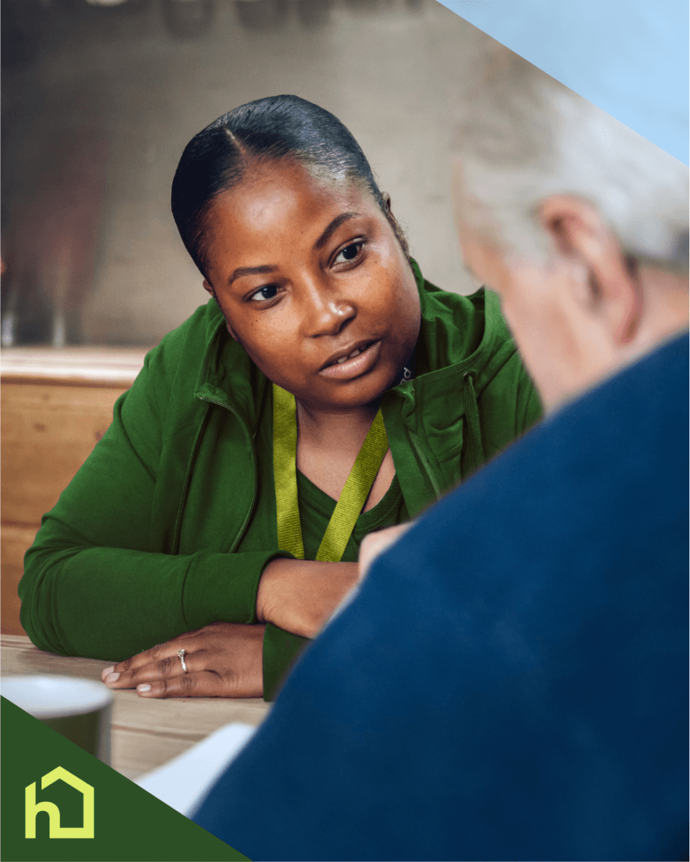 A woman in green listens attentively to an older man across a table, with a house logo in the corner. - Home Instead