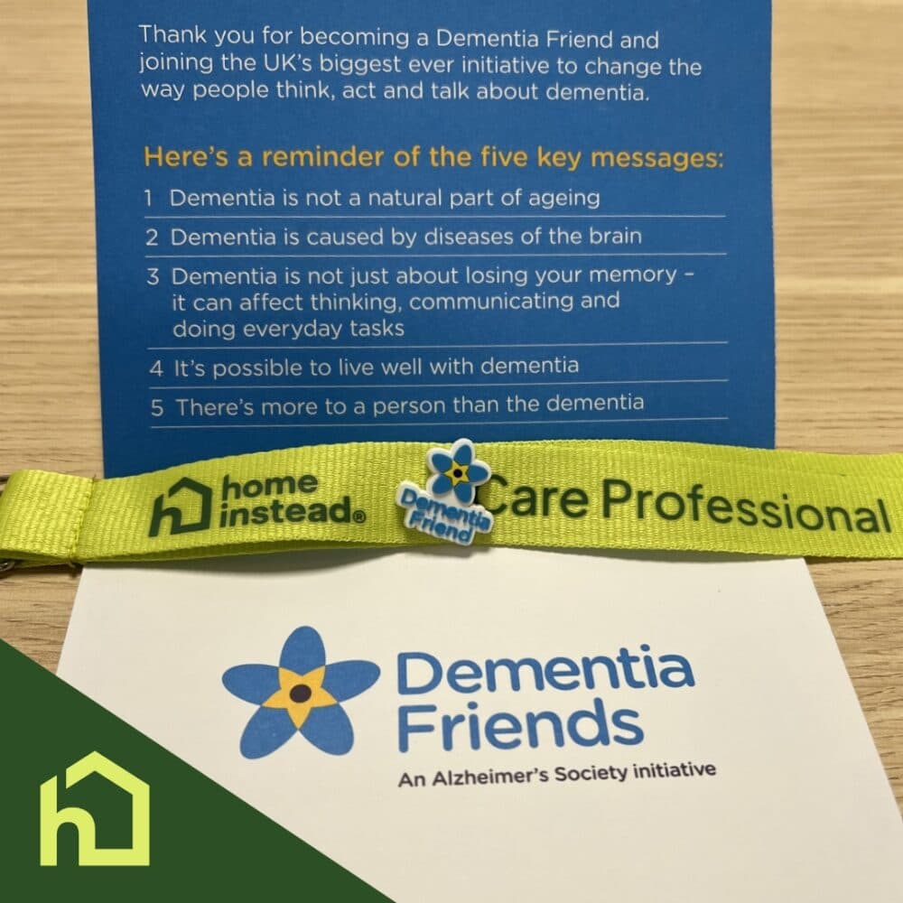 Dementia Friends info card, yellow ribbon, and Dementia Friends badge on a wooden table. - Home Instead
