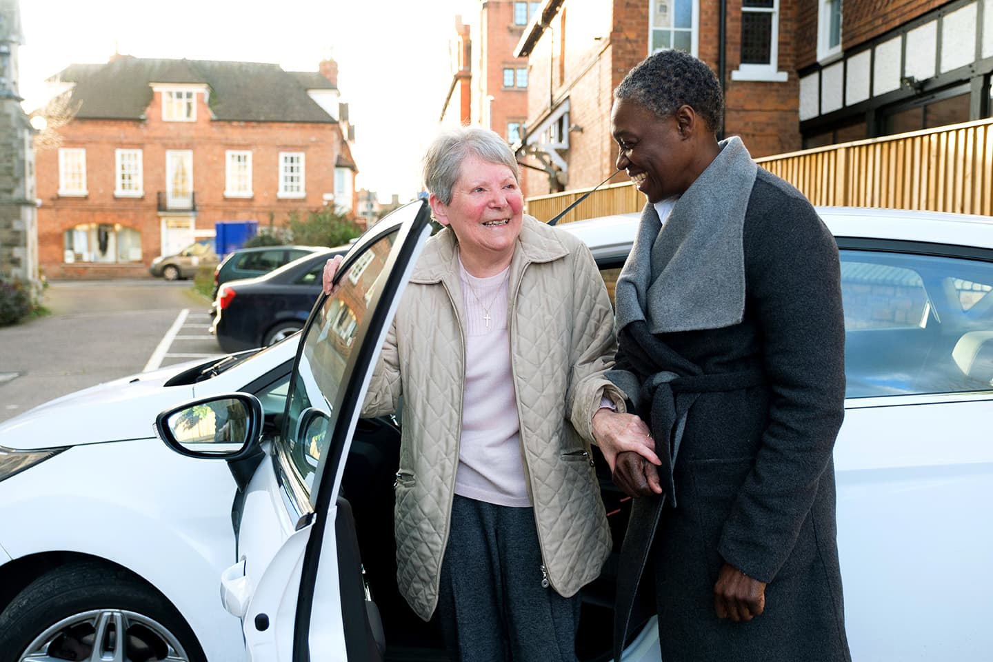 Two women smile together by a white car in a parking lot, one holding the car door open for the other. - Home Instead