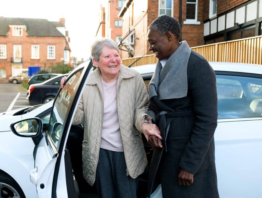 Two women smile as one helps the other out of a car in a residential neighborhood. - Home Instead