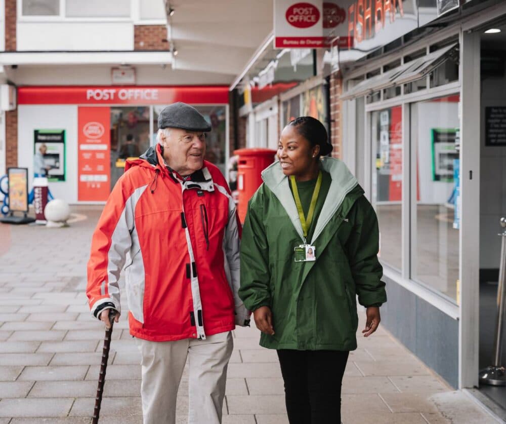 An elderly man with a cane walks beside a smiling caregiver outside a post office. - Home Instead