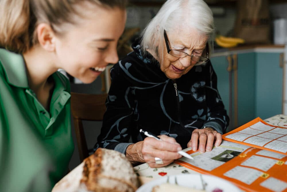 Elderly woman filling out a calendar with a pen while a younger woman sits and smiles beside her. - Home Instead