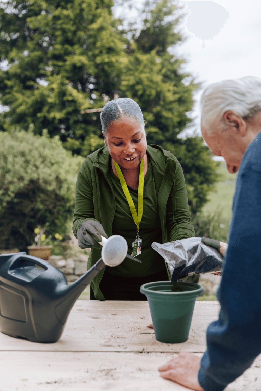 A woman and an older man potting a plant together outdoors, smiling, with gardening tools on the table. - Home Instead
