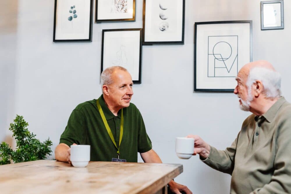 Two older men sit at a wooden table, holding white mugs and talking, with framed art on the wall behind them. - Home Instead