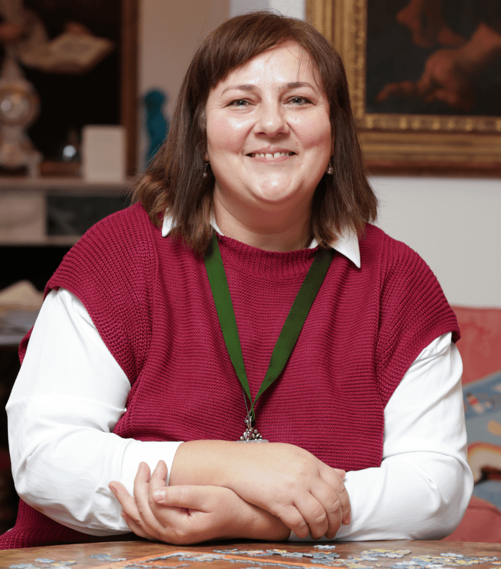 A woman in a red sweater and white shirt smiles while sitting at a table with a puzzle. - Home Instead